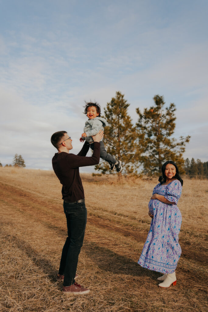 Dad tossing toddler in the air with mom's hands on her bump looking at the camera