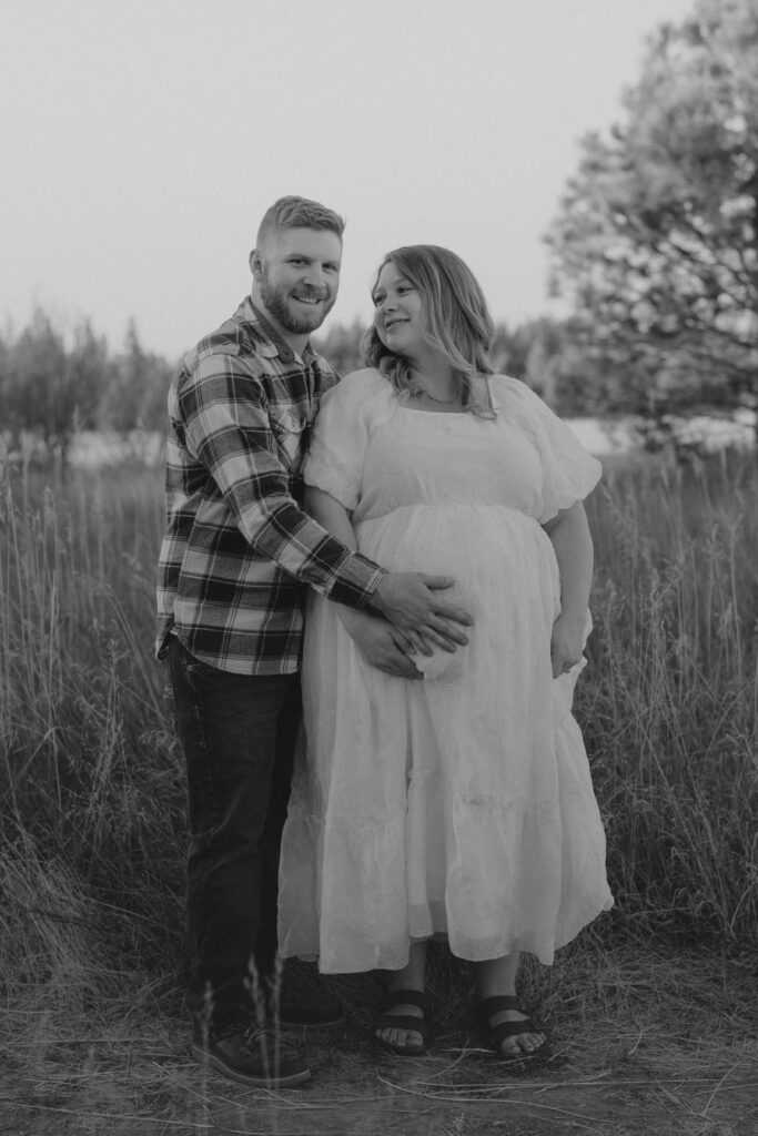 when to schedule maternity photos - husband with hand on wife's baby bump in field, black and white