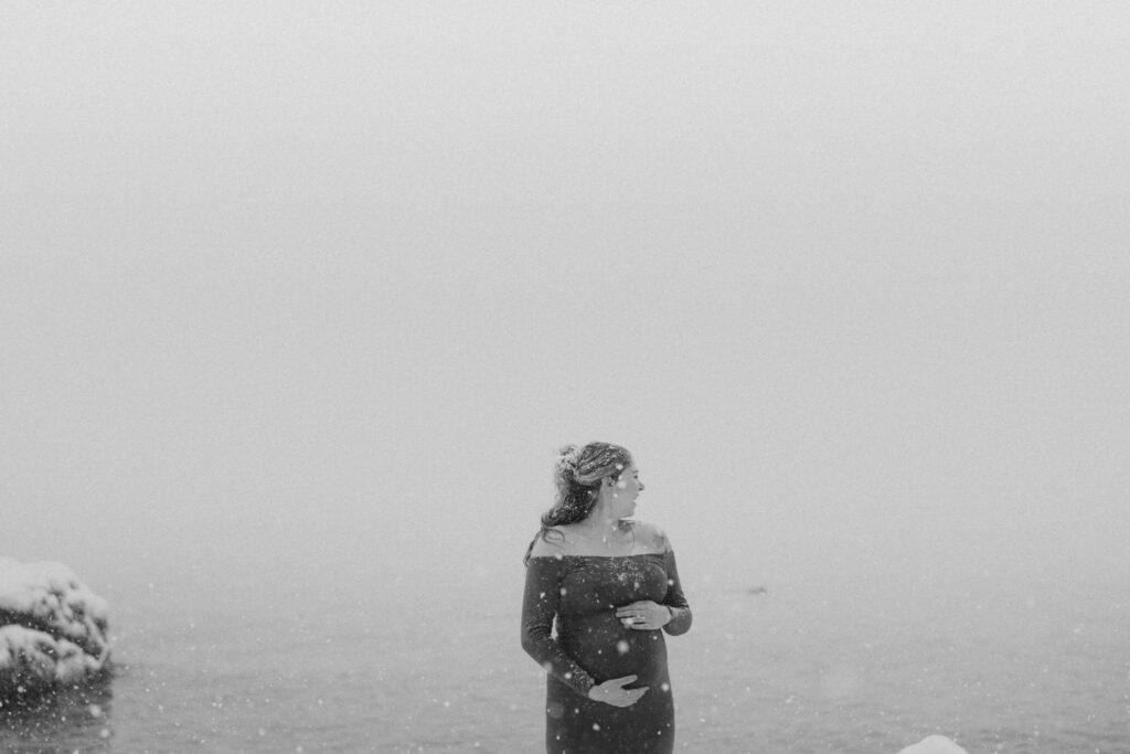 Black and white of woman in snow with her hands on her bump looking over he shoulder
